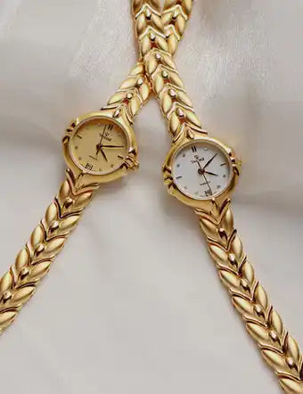 Two elegant women’s gold-toned watches with decorative link bracelets, one with a cream dial and Roman numerals, the other with a white dial and stick markers.